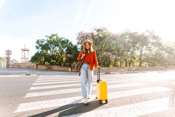 Woman stands at crosswalk with yellow suitcase next to her. A beautiful tourist in hat and backpack crosses the road with a brightly colored suitcase. Concept is tourism, style, and urban. © maxbelchenko