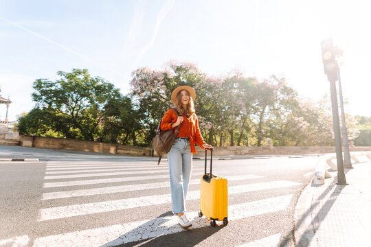 Woman stands at crosswalk with yellow suitcase next to her. A beautiful tourist in hat and backpack crosses the road with a brightly colored suitcase. Concept is tourism, style, and urban.
