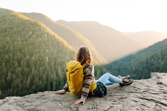 A female hiker with a backpack sits on the edge of a cliff at sunset. The young woman enjoys the mountain scenery outdoors. Concept of relaxation, hiking, and nature.
