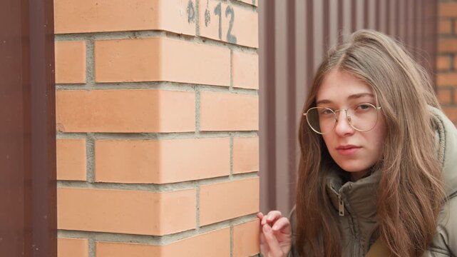 Young woman leaning against brick wall closeup, puffer jacket and glasses, thoughtful gaze and quiet expression, textured masonry backdrop, soft snowfall and intimate contemplative atmosphere