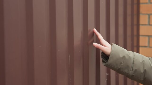 Caucasian woman touches brown metal fence near brick pillar, winter coat and glasses, homebuyer inspecting property boundary with cautious fingertips, closeup texture study, muted overcast street
