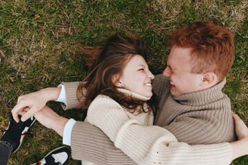 Obraz premium A young couple embraces and smiles warmly while lying on the grass wearing cozy sweaters in a natural outdoor setting, capturing a candid moment of connection and happiness.