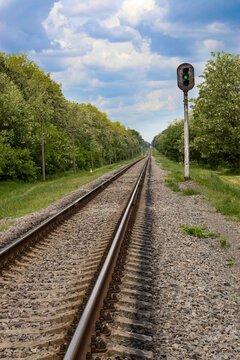 Train tracks run straight through an open area with trees lining both sides. A green signal light stands by the tracks. The sky is blue with some clouds.