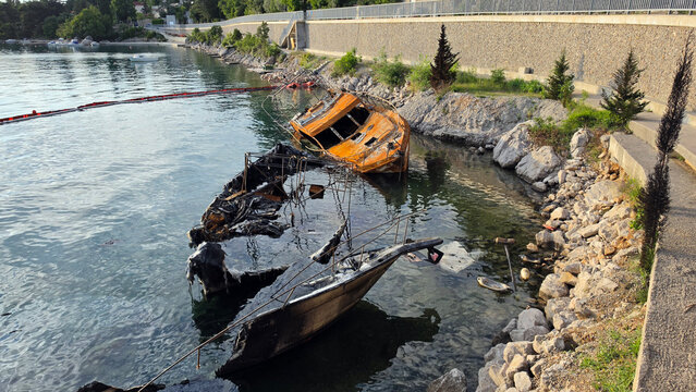 Two heavily burned and destroyed motorboat shipwrecks partially submerged in shallow water near a rocky coast retaining wall