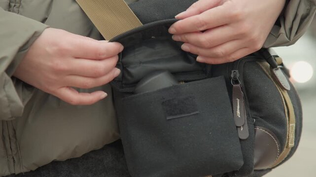 Hands tucking item into canvas pouch on hip, closeup of careful placement and soft flap closure, khaki strap and muted city lights in background, scene conveys organization and small gear care