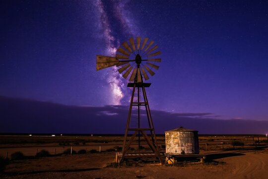 Old windmill stands against blazing milkyway in karoo night. Farm water pump on dirt ground. Vast desert landscape under stars. Renewable energy and ancient tech.