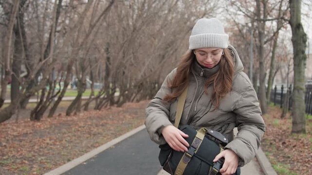 Woman walking in park searching bag for camera lens, wearing beanie and puffer coat, overcast autumn day, bare trees lining pathway, candid pause as commuter and student checks messenger bag.