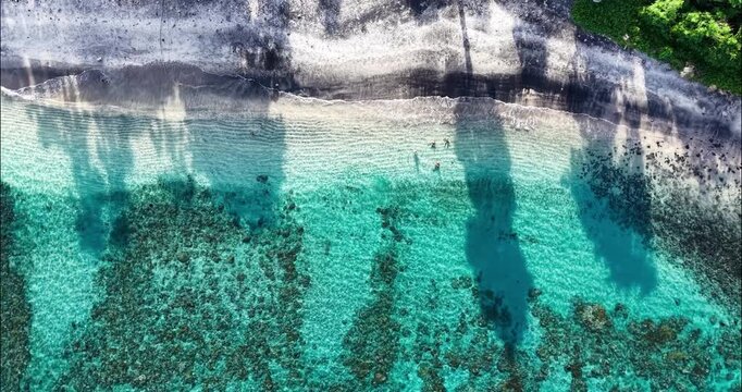 Overhead aerial drone shot of crystal-clear turquoise water, coral reef formations, and a volcanic black sand beach in Amed, Bali, Indonesia with swimmers enjoying the tropical shoreline.