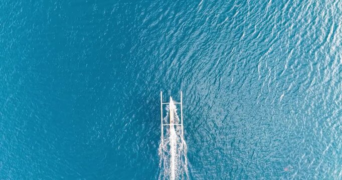Overhead aerial drone shot of a traditional Balinese jukung boat creating a wake across deep blue tropical ocean waters near Amed, Bali, Indonesia.