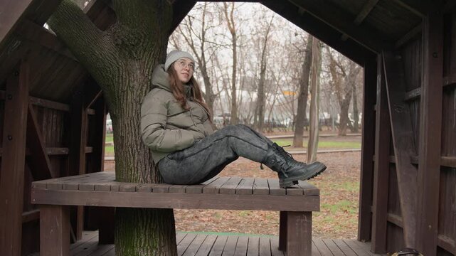 Young woman resting under wooden shelter, sitting against tree trunk on bench, wearing knit beanie, puffer jacket and boots backpack at feet relaxed upward gaze and reflective mood autumn leaves