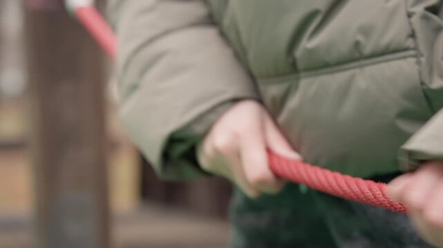 Closeup hands gripping red rope framed by puffy green jacket, knuckles tensed, slow pull across playground post, autumn light, tactile rope strands, intimate study of effort and focus, cinematic