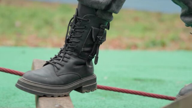 Combat boot stepping on rope plank above green mat, confident forward stride and heavy tread pressing into wood, worn leather and laces in motion, determined momentum and outdoor training vibe