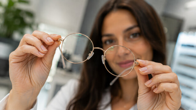 Woman holding round eyeglasses while choosing frames in an optical store. Concept of vision correction, ophthalmology, eye care and eyewear selection.