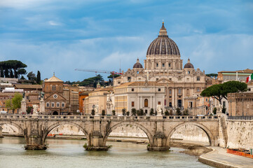 Fototapeta premium View on stone-arched bridge on Tiber river and St Peter's Basilica in Rome, cloudy sky, ochre and terracotta buildings, domes and umbrella pines rise on gently sloping Vatican hill