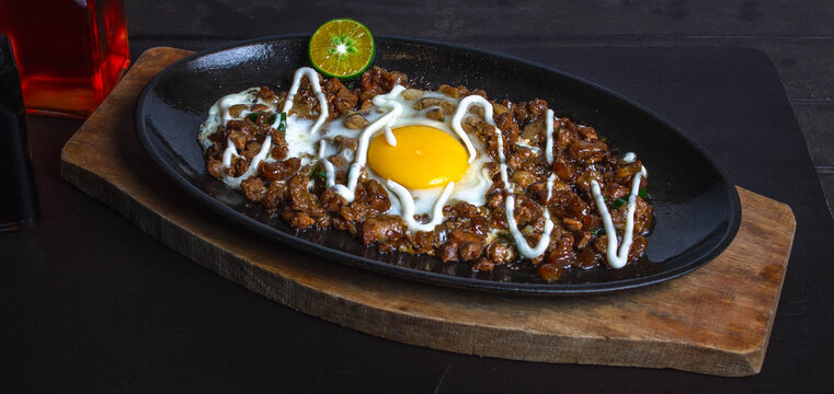 close up photo of spicy pork mask sisig on a sizzling plate with egg and lemon drizzled with mayonnaise.