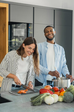 Happy diverse couple cooking together in a bright modern kitchen, woman chopping colorful vegetables while man stirs a pot, smiling and enjoying healthy home meal preparation