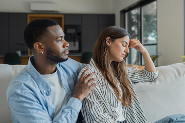 Fototapeta premium Young couple sitting on a sofa at home, the man offering comfort to the sad woman struggling with emotional distress after a relationship disagreement