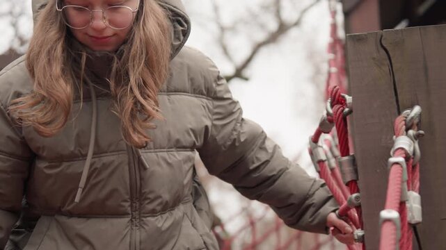 Cautious white girl crossing rope bridge in playground, puffer jacket and beanie, steady grip on ropes, careful footing and focused gaze, youthful explorer negotiating timber structure under overcast