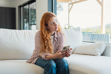 Obraz premium Red-haired woman sitting on a white sofa, holding a smartphone and looking away thoughtfully, relaxing in a bright modern home living room with large windows