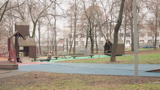 Empty playground wooden playset autumn morning, brown leaves scattered on blue safety surface, leafless trees and distant apartment buildings, quiet park atmosphere, municipal equipment and worn