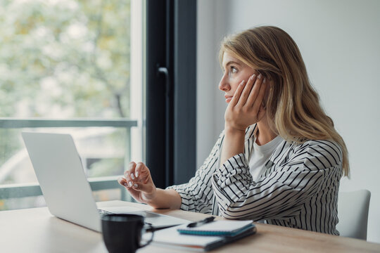 Woman sitting at a desk and looking out the window, distracted from work, showing concepts of procrastination, boredom, thinking, and lack of motivation during remote tasks