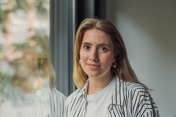 Fototapeta premium Young blonde woman smiling, wearing a striped shirt and gold hoop earrings, looking at the camera while standing by a window with natural light creating reflections