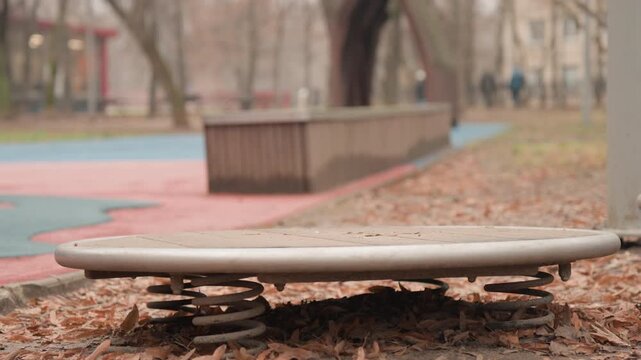 Single boot hopping on round spring platform, playground coil compresses and rebounds while person explores motion and rhythm, vibrant yet muted urban park setting with benches and onlookers blurred