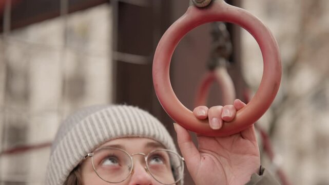 Reaching white woman gripping playground ring, focused gaze through glass frames, fingers wrapping around weathered metal, steady upward motion, tension and determination, urban winter backdrop