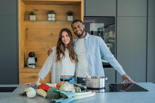 Joyful multi ethnic couple standing in a stylish kitchen embracing while preparing a healthy meal with fresh vegetables and cooking a dish on the stovetop