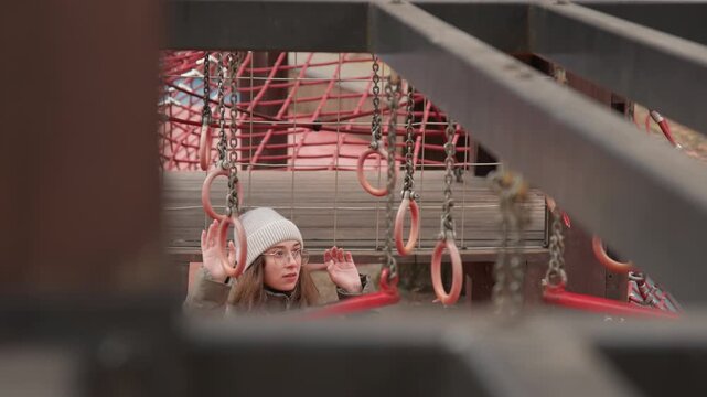 Young girl exploring playground rings winter, bundled knit hat and coat, peeking through metal beams, ropes and chains visible, playful curiosity meets quiet snowy atmosphere, closeup on expression