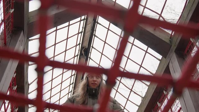 White woman on playground net looking down through red grid, wearing winter coat and beanie, overhead metal frame visible, careful posture and measured steps, testing balance, documentary mood, high