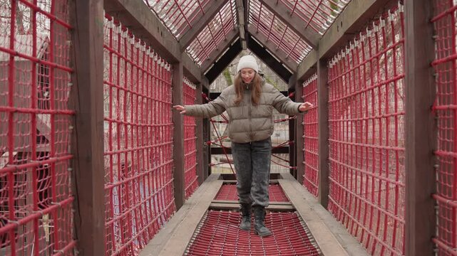 Fullframe white woman balancing on red lattice walkway, bundled in puffer coat and knit hat, playful winter stroll with hands on rails for stability, warm smile and festive architectural corridor