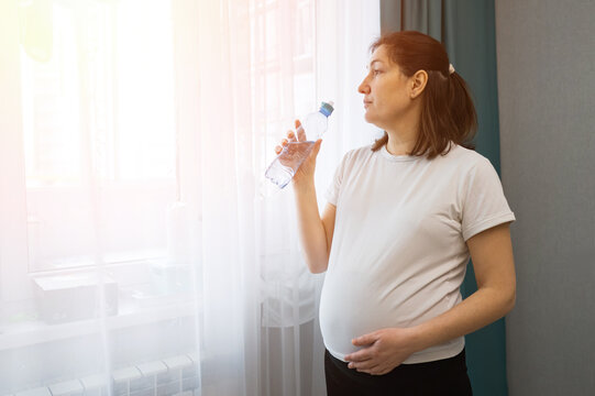 Pregnant woman standing by a bright window and drinking water from a bottle, home wellness scene with sheer curtains, teal drapes, and soft daylight