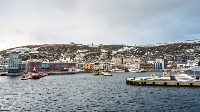 Beautiful view of boats and buildings along the waterfront of Hammerfest in Norway with snow-covered hills in the background during a cloudy day