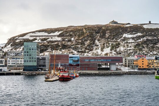 Beautiful view of boats and buildings along the waterfront of Hammerfest in Norway with snow-covered hills in the background during a cloudy day