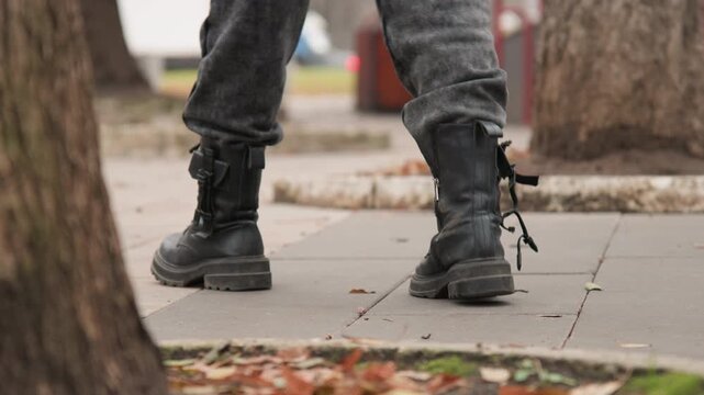 Black combat boots walking on sidewalk, lowangle closeup of laced leather boot stepping across concrete pavement with scattered autumn leaves, tree trunk edge in foreground, cool overcast light,