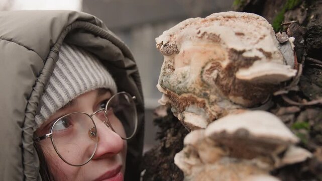 Woman examines fungus. Person observes polypore fungus on mosscovered tree trunk carefully. Closeup of woman in winter clothing analyzing fungal resource on tree bark with gentle smile