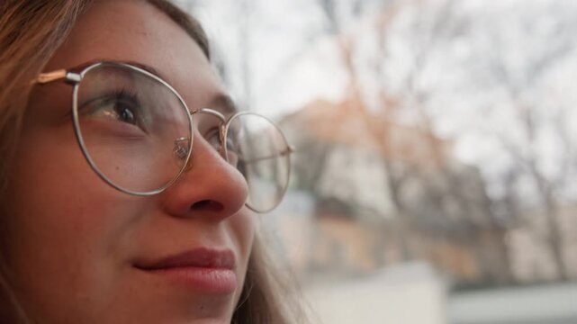 Caucasian woman smiling under bare trees, soft golden light, glasses catching highlights, gentle breeze, cozy sweater tones, hopeful gaze across city square, serene autumn atmosphere