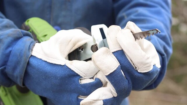 A worker in protective gloves replaces the blade on a sabre saw. A craftsman in a blue work uniform and strong protective gloves installs a saw blade into the chuck of an electric sabre saw