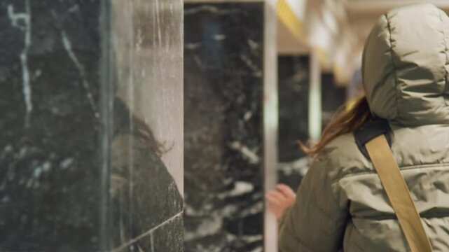 Hooded traveler brushes marble pillar, hand lingering on cool stone, long hair peeking from hood, subtle glance around corridor, tactile exploration of public space, hesitant rhythm and reflective