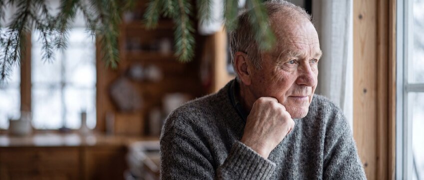 Senior man looking out window in cozy cabin thinking. Elderly thoughtful male in grey sweater contemplating life by glass pane. Pensive mature adult in retirement home feeling lonely and nostalgic.