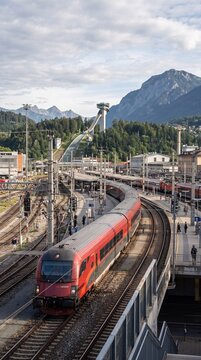 An aerial view of an &Ouml;BB Railjet passenger train departing the Innsbruck train station in Austria, with the iconic Bergisel Ski Jump visible in the mountains.
