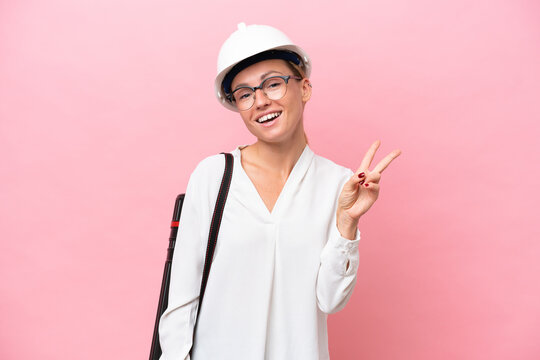Young architect Russian woman with helmet and holding blueprints isolated on pink background smiling and showing victory sign