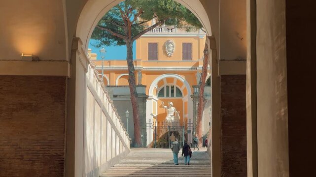 Tourists explore stunning historic buildings and courtyards in Rome, Italy, featuring grand classical architecture, framed archways, statues, and palm trees under a bright blue sky during the daytime.