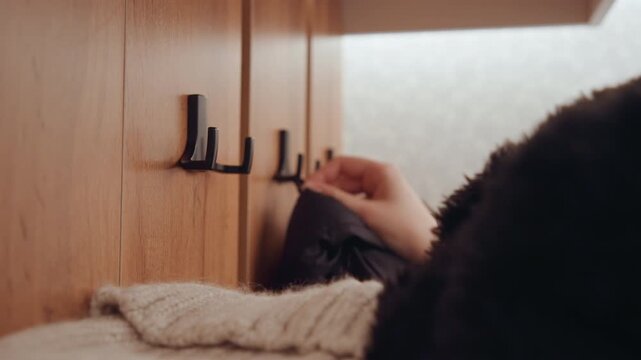 Closeup coat hook with jacket being picked up, gentle hand motion, knitted scarf and sweater in foreground, soft warm tones, subtle pause before departure, hushed domestic mood, tidy entry space