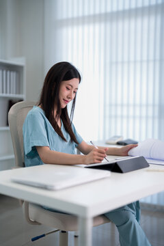Asian young adult female doctor healthcare professional writing patient medical report notes at hospital office desk wearing scrubs working on documentation
