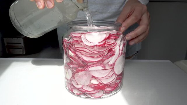 Person pouring brine into glass jar filled with sliced radishes for pickling. Symbolizes healthy eating, food preservation, and fermented vegetables. Ideal for nutrition, culinary and wellness content
