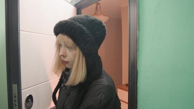 Blonde woman stands at door pensive, lips pursed and gaze lowered in narrow green corridor, black beanie and quilted jacket, soft lamp glow and quiet apartment landing creating intimate pause