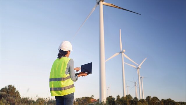 Young technical professional checking data on a computer, standing at a wind power plant against a bright blue sky, engaged in an eco-friendly energy project