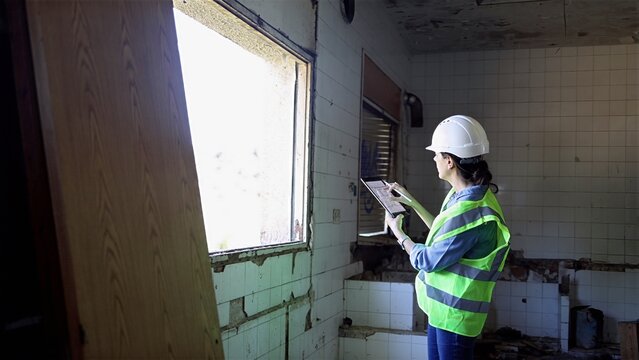 Woman wearing hard hat and reflective vest documenting structural issues and surveying a severely damaged room with a tablet, planning for restoration, rehabilitation, or demolition